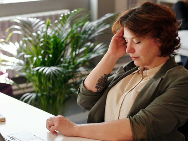 Woman in office setting, sits at a desk and cradles her head, indicating pain. There is a fern plant in the background.></a></div><div class=