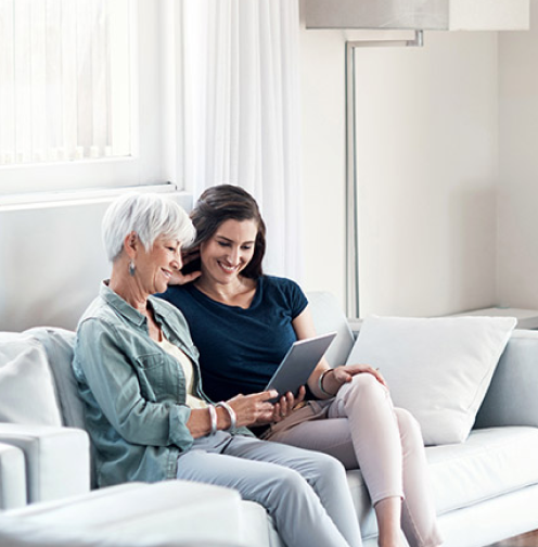 News - An older woman with white hair and her daughter sitting together on a couch looking at a tablet