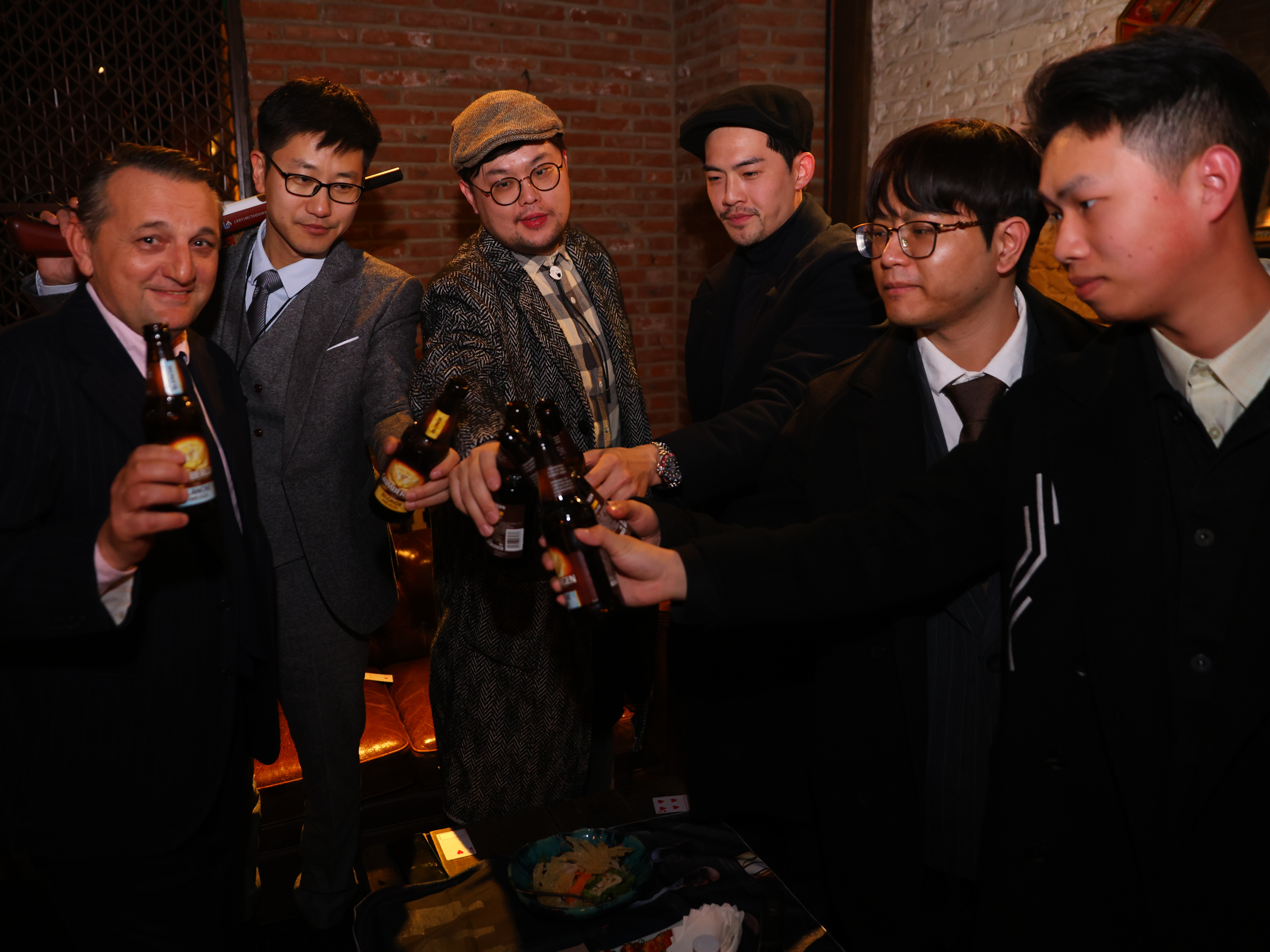 Picture of a group of six men in different colored suits toasting with a beer.