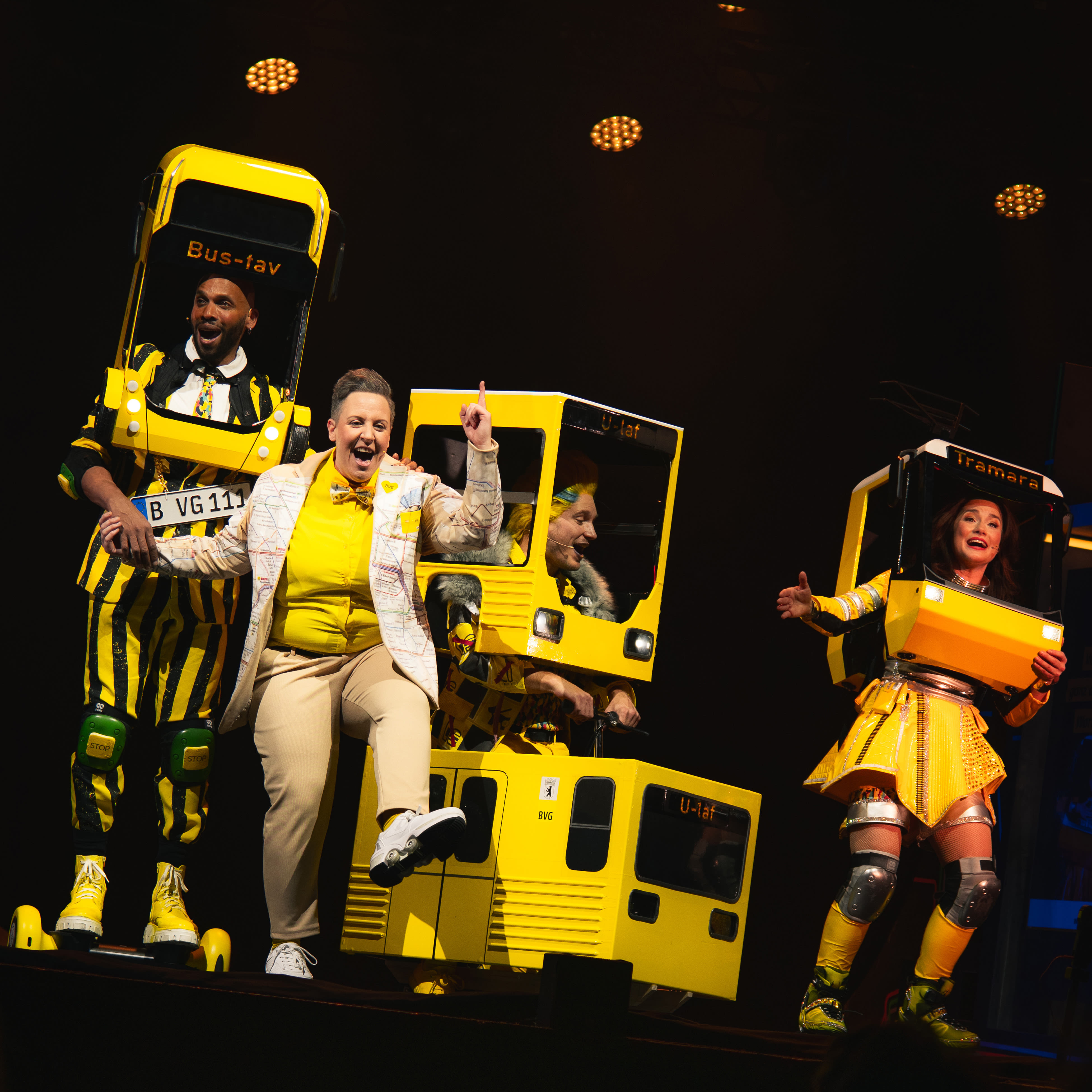 Performers on stage in vibrant costumes with theatrical props, depicting a scene involving a yellow bus and a phone booth, with dynamic lighting effects enhancing the ambience of the performance.