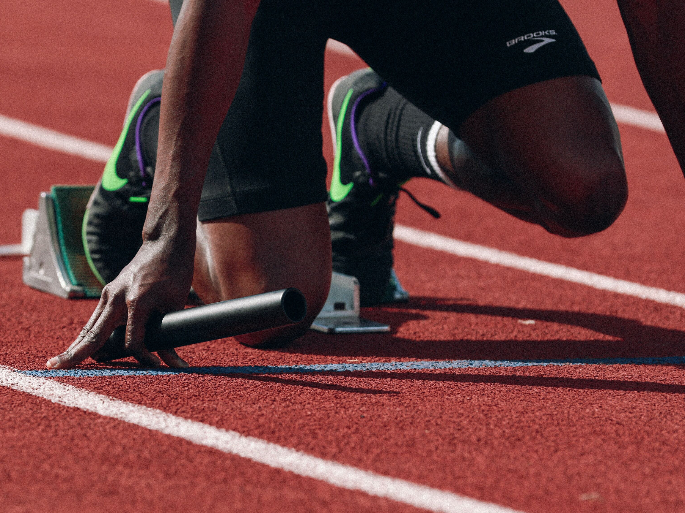Image of the lower body of a relay runner in starting position on a running track. On the pants you can see the brand Brooks.
