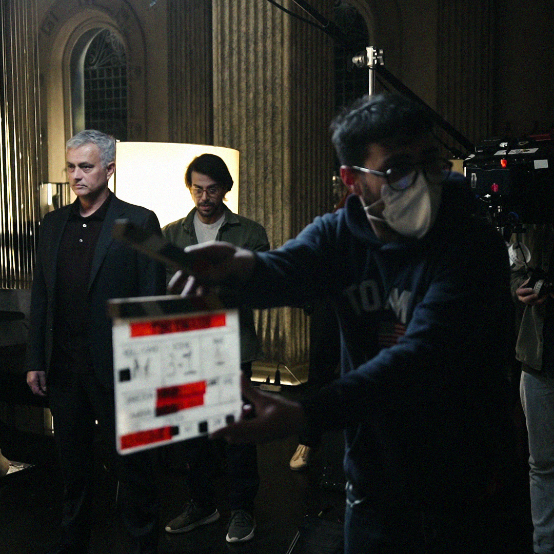 Picture from the shoot: A dark room with two columns on the left. about 10 people, some wearing masks, stand around camera equipment. a large light is held in from the side. Mourinho in the middle from the back.