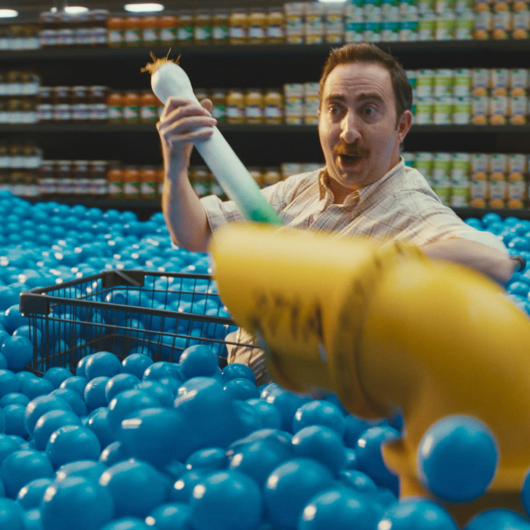 A man with dark hair and a light shirt is seated among a pile of blue plastic balls in a grocery store. He holds a large piece of yellow plumbing pipe and has a shopping basket beside him, with shelves of canned goods visible in the background.