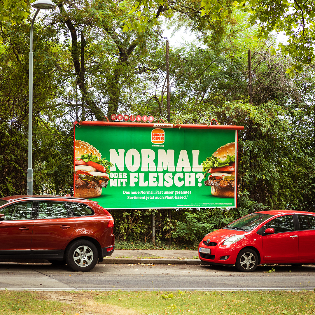 Picture of streets with green strip in the middle. Several cars parked at the roadside. In the background three posters of Burger King advertising "regular or meat-based?