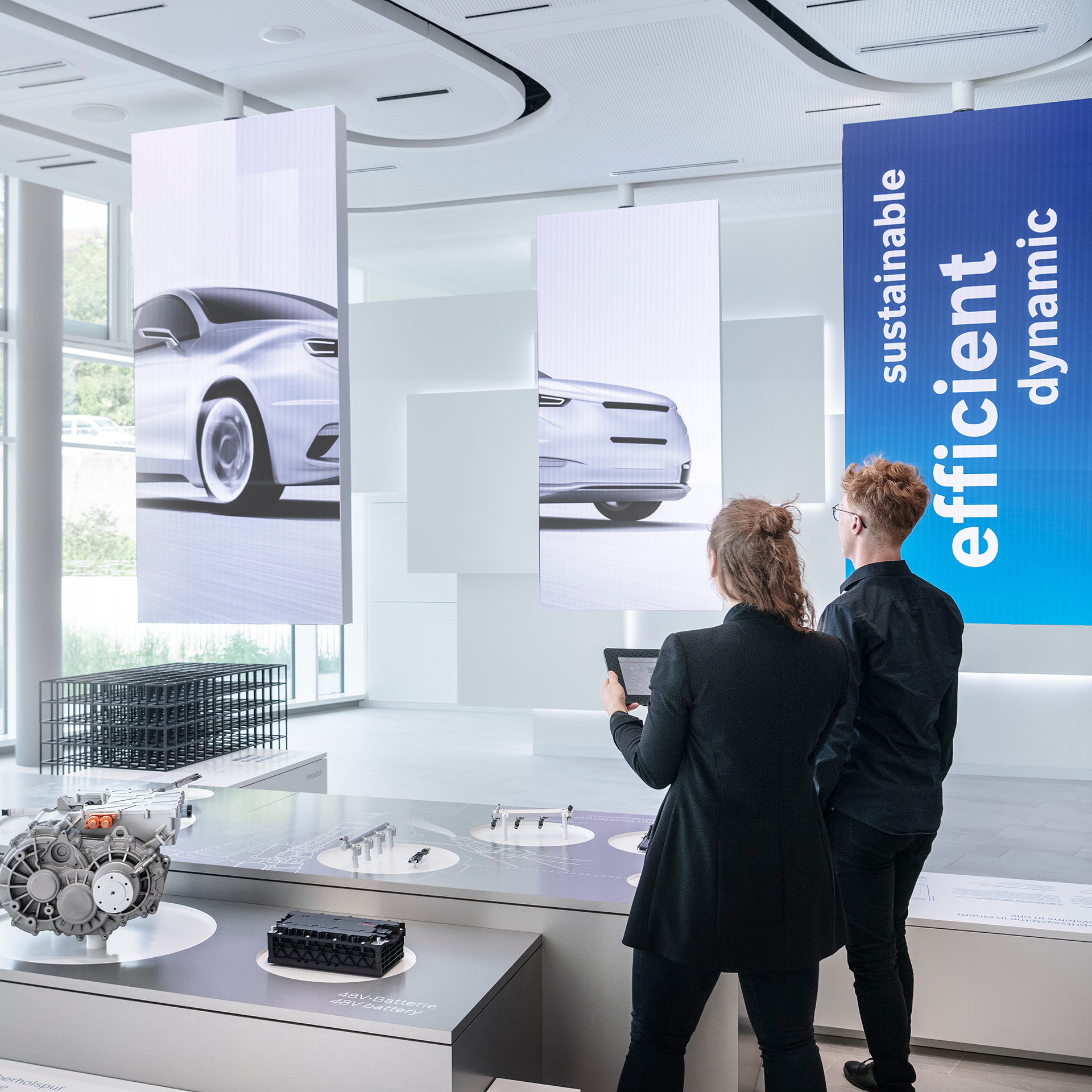 Two people admiring a sleek car in a bright showroom, surrounded by other vehicles and modern decor.