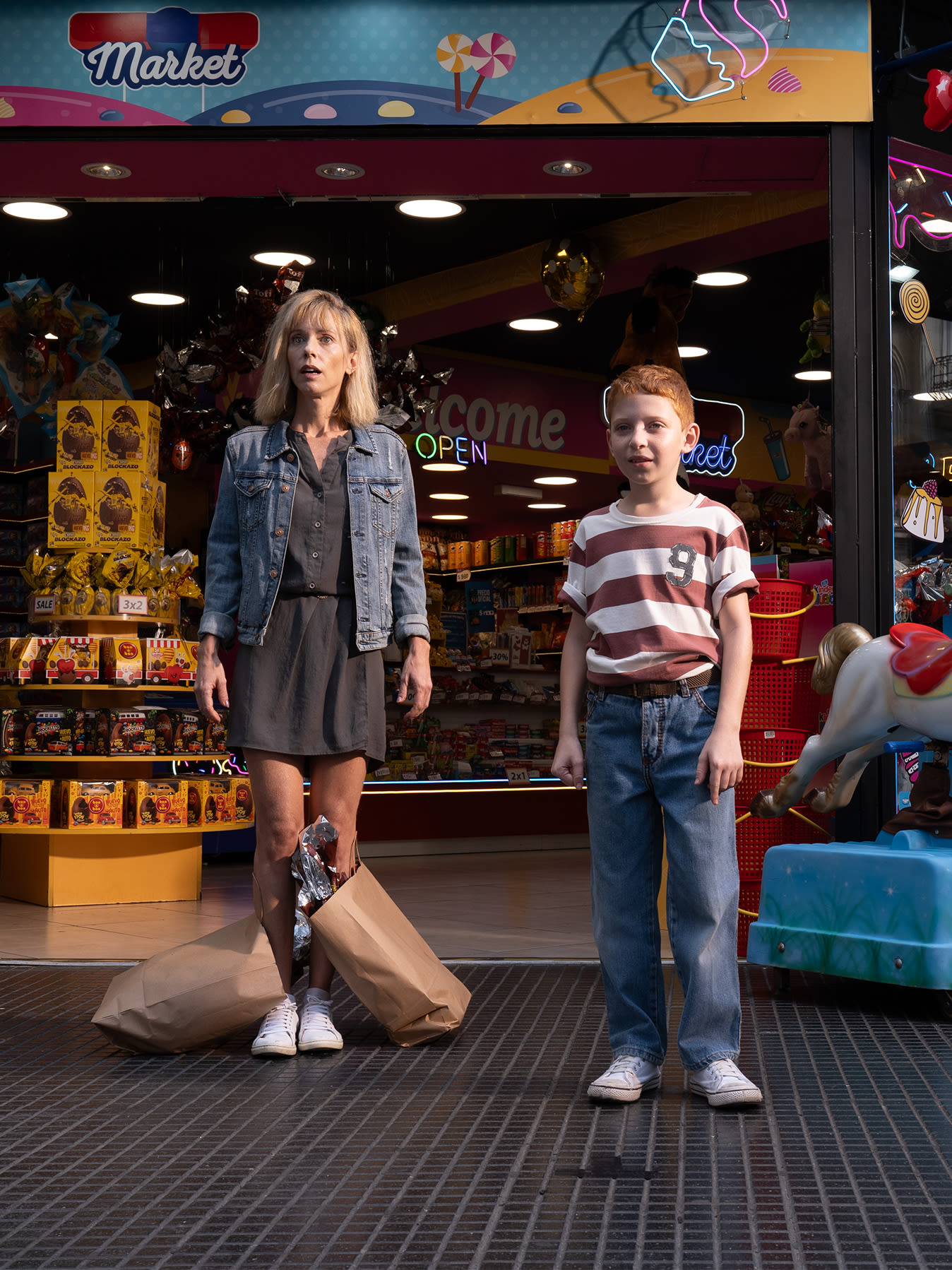 A woman and a boy smiling together in front of a colorful store entrance, ready for a fun shopping trip. 