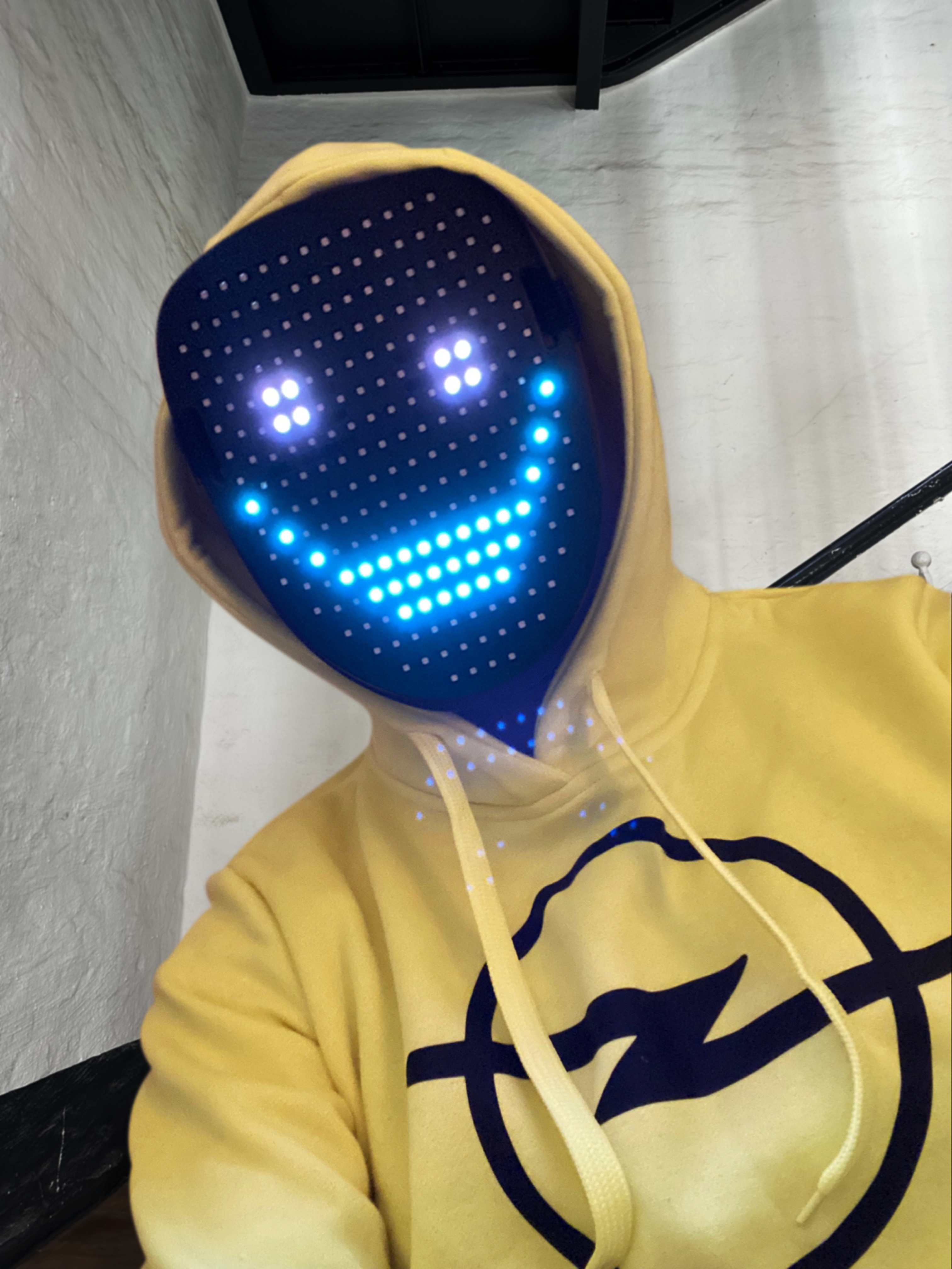 Picture of a person in a yellow hoodie with Opel logo on it. Above the face is a LED mask that represents a grinning face through points of light.