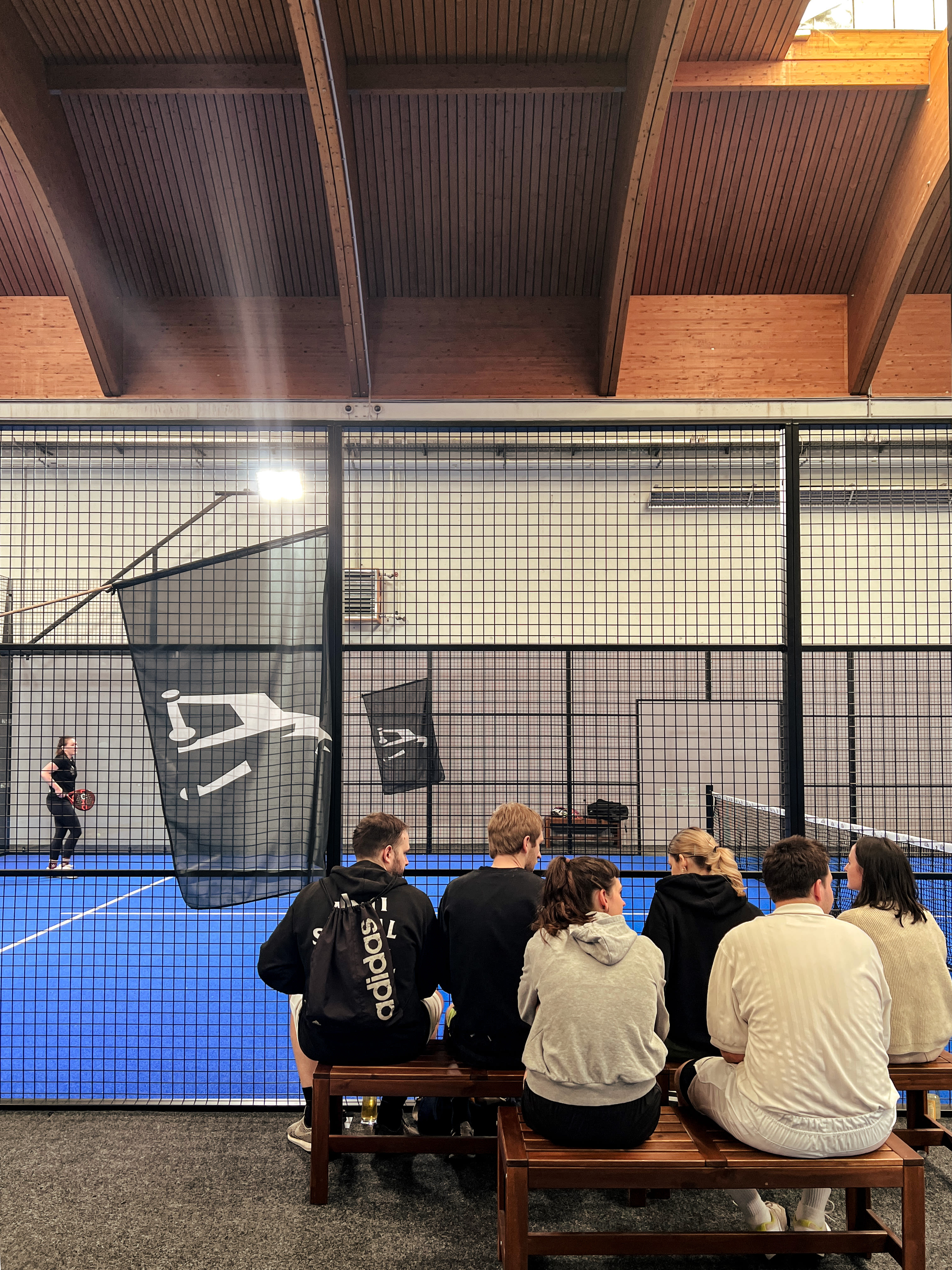 Image of a group of Jung von Matt DONAU employees sitting in front of a cage court of the trend sport Padel watching others.