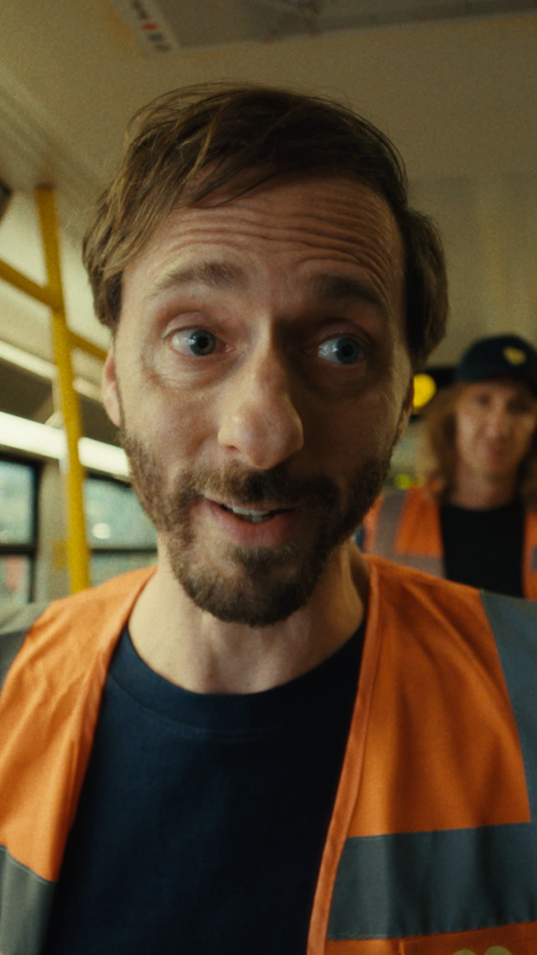  A man wearing an orange vest and safety gear sits on a subway train, looking relaxed and ready for his commute.