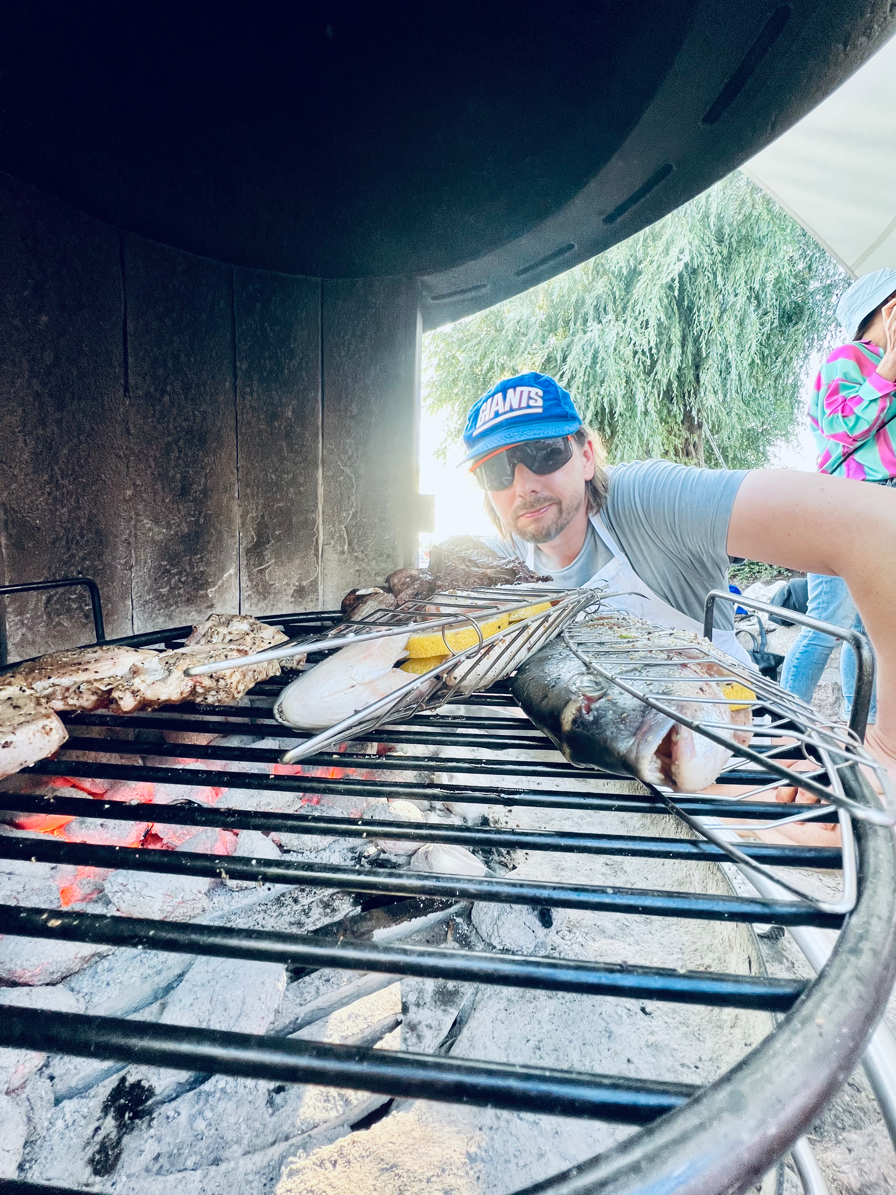 Image of an employee with sunglasses and cap from Jung von Matt SPREE posing in front of a charcoal grill on which two fish are being grilled.