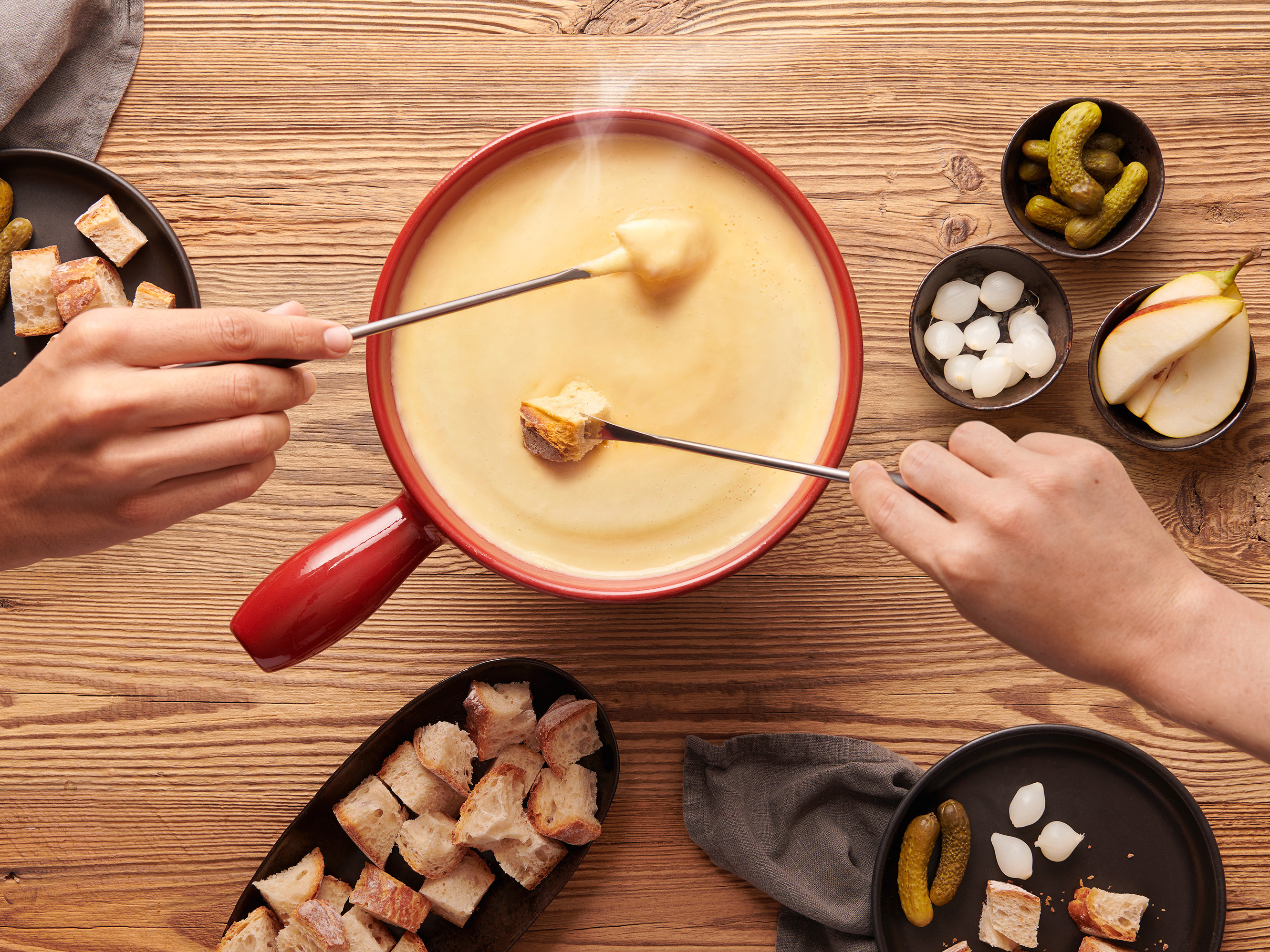 Picture of a cheese fondue from above, standing on a wooden floor. Two hands with forks dip various ingredients in it.