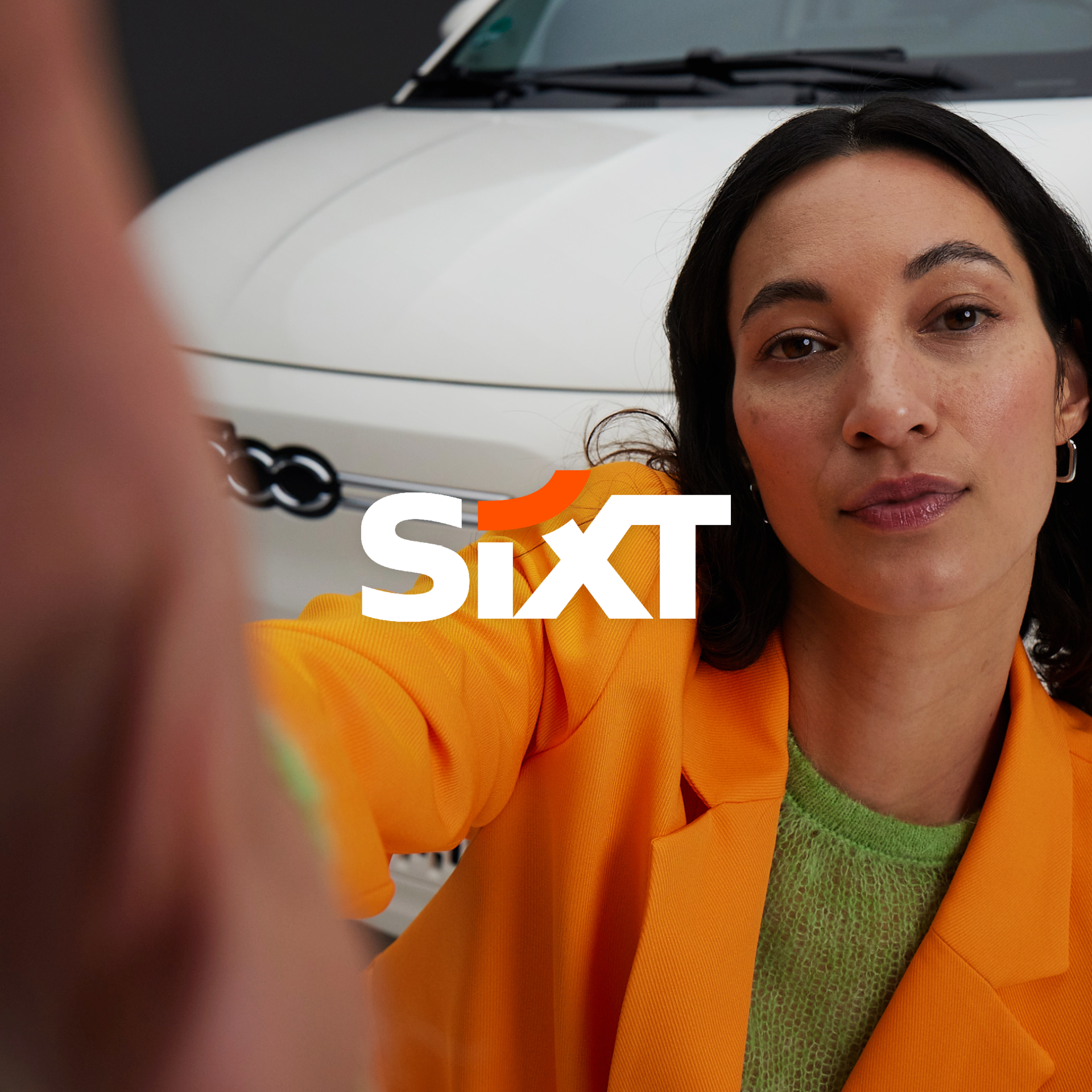 SIXT ad in new brand design by Jung von Matt BRAND IDENTITY: a young women with a blazer jacket in SIXT Orange standing in front of a white FIAT 500 making a selfie with her smartphone, the hand a bit in front of the camera