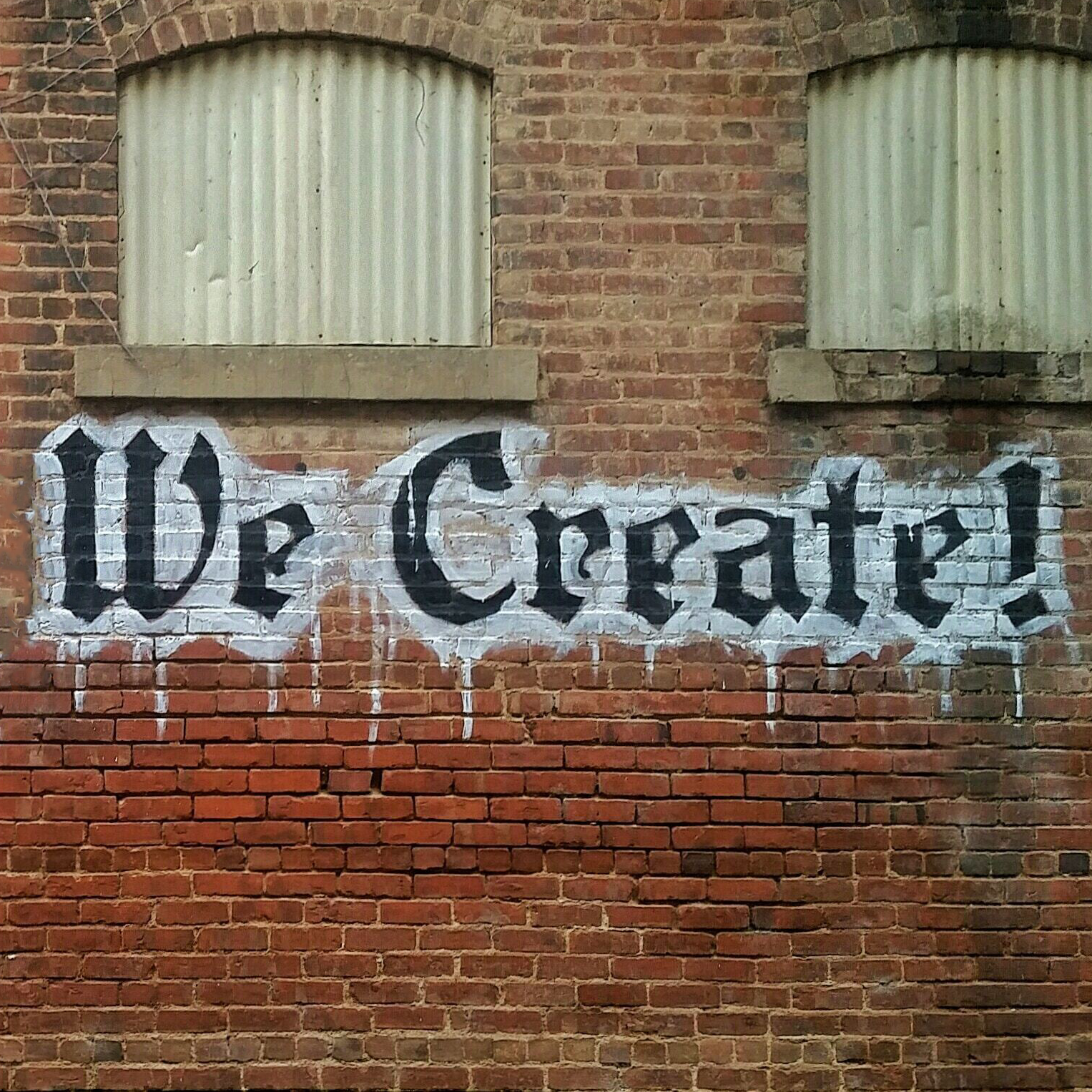Image of a red brick wall. On the wall is written in black and white: "We Create".