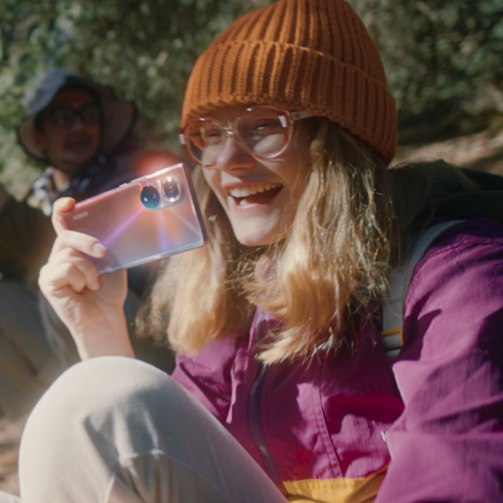 Image of a smiling woman in hiking gear taking a photo with an Honor 50 smartphone. In the background, an out-of-focus male person with a hiking stick.