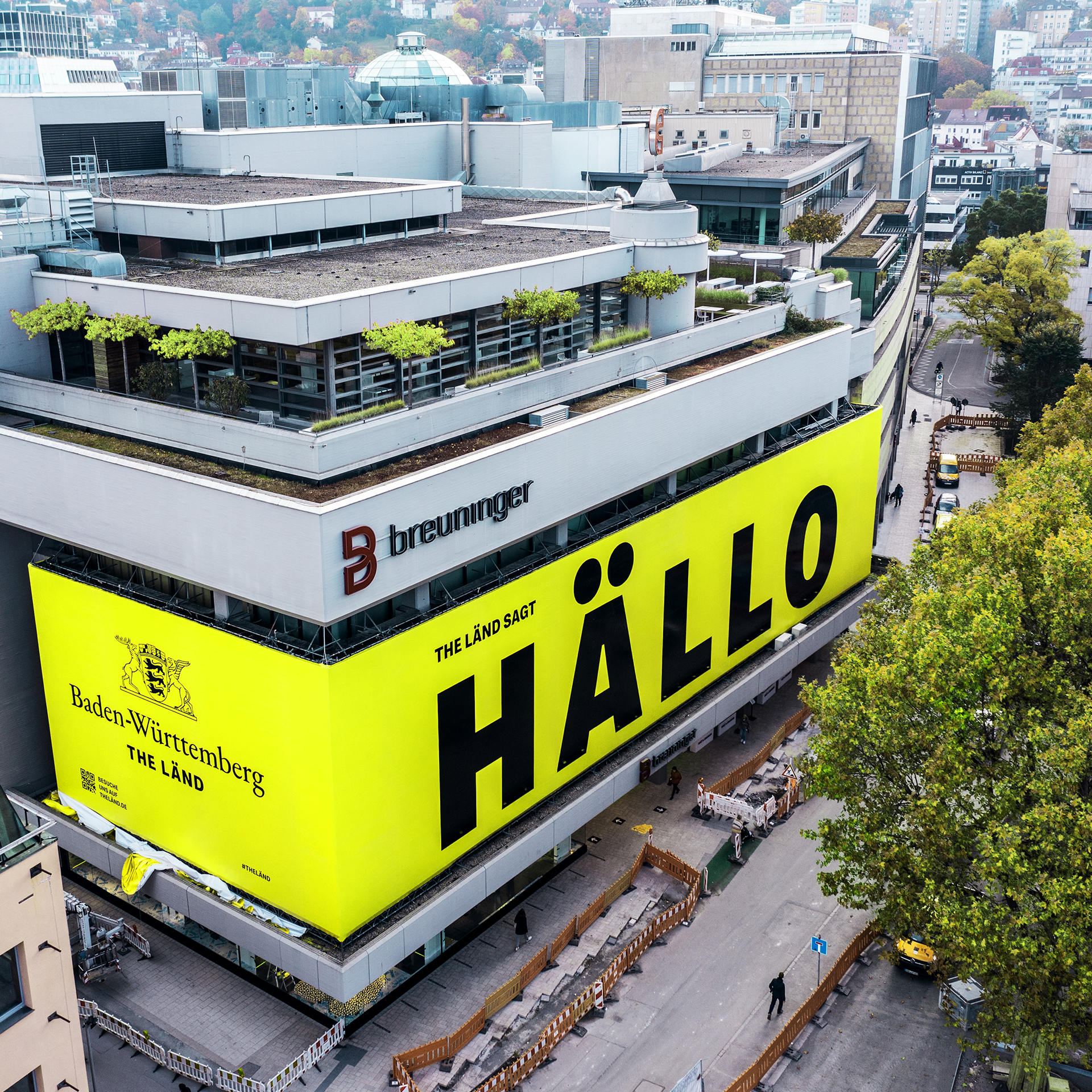 Aerial view of a departement store building with the lettering Breuninger. All around is a large yellow poster that says The Land sagt Hällo.
