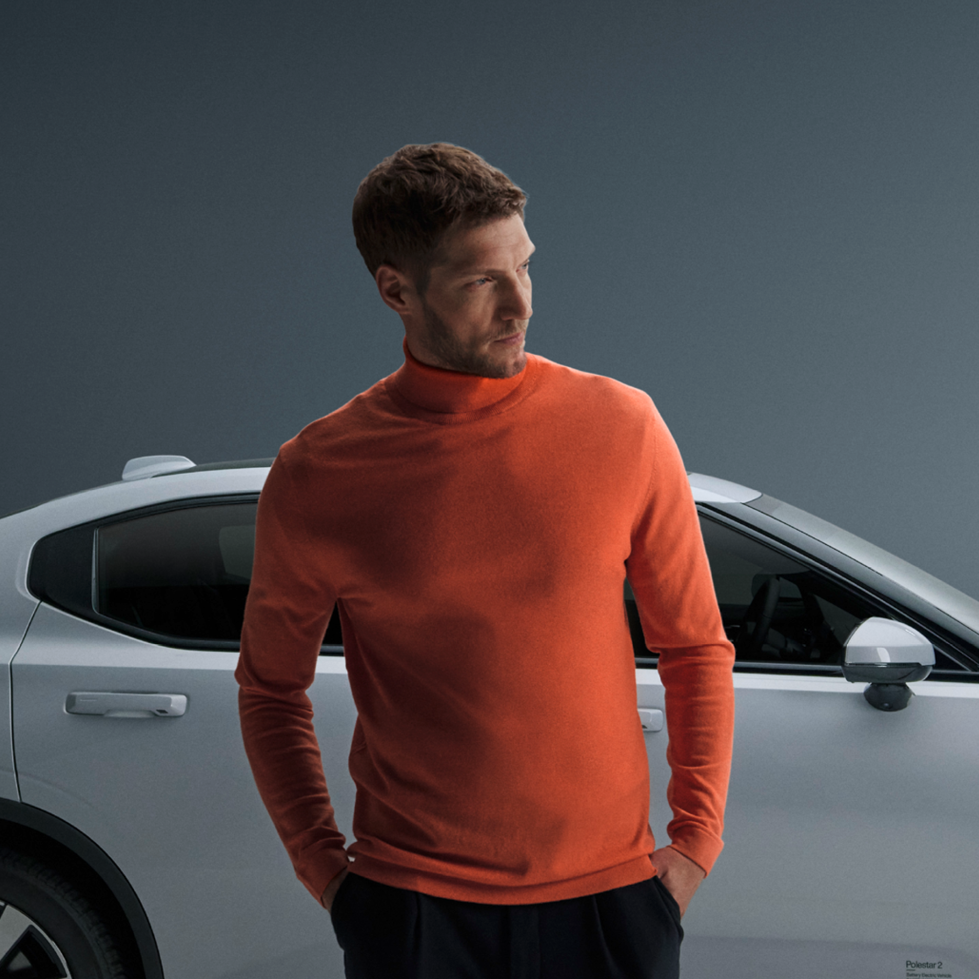 Male person with a pullover in SIXT ORANGE standing in front of a white Polestar electro car looking sideways, new brand design by Jung von Matt BRAND IDENTITY for iconic car rental company SIXT