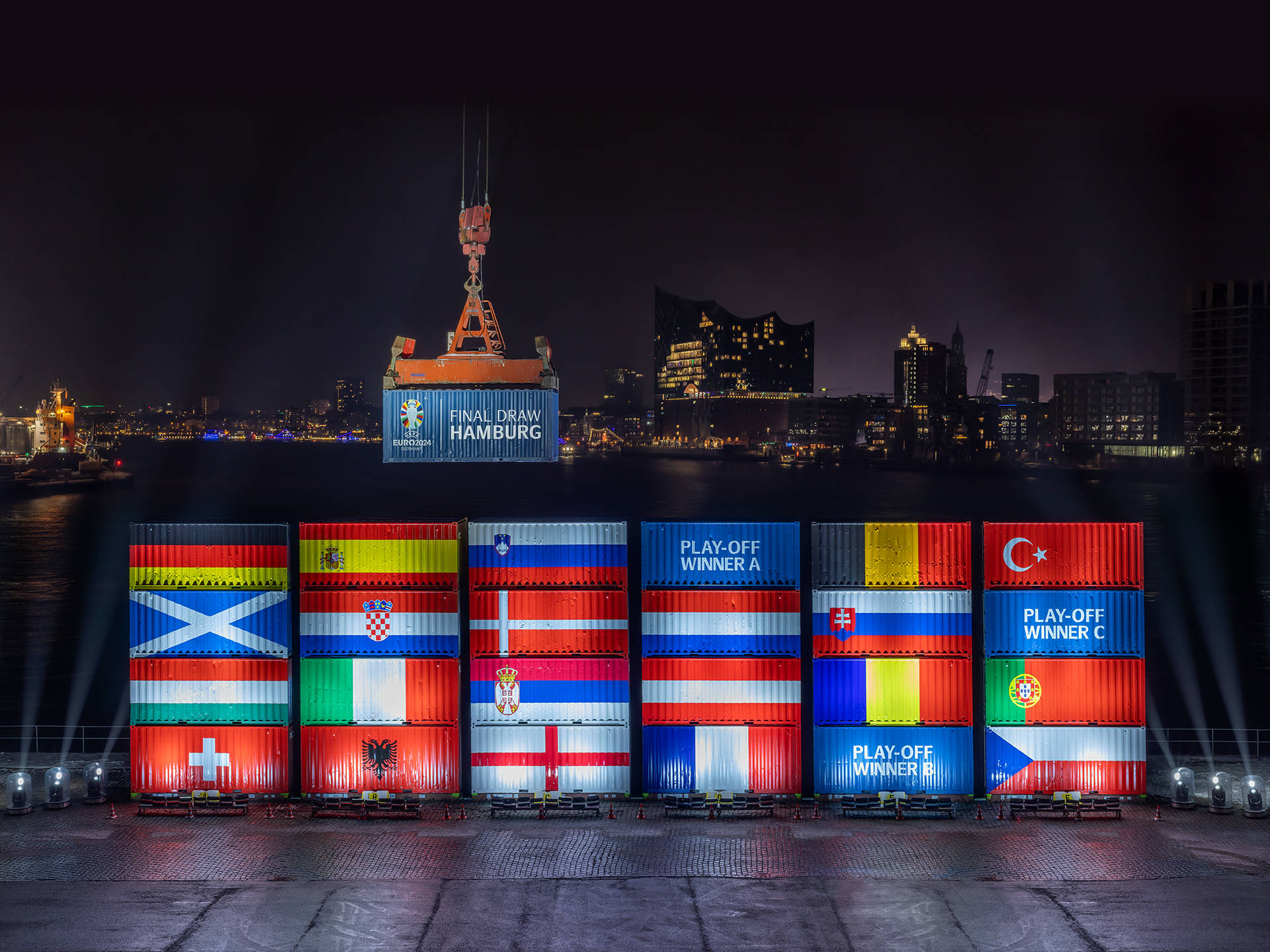 A nighttime view of large shipping containers, displaying various national flags and labeled "Play-Off Winner." Behind them, the Hamburg skyline and cranes are visible under dark skies.