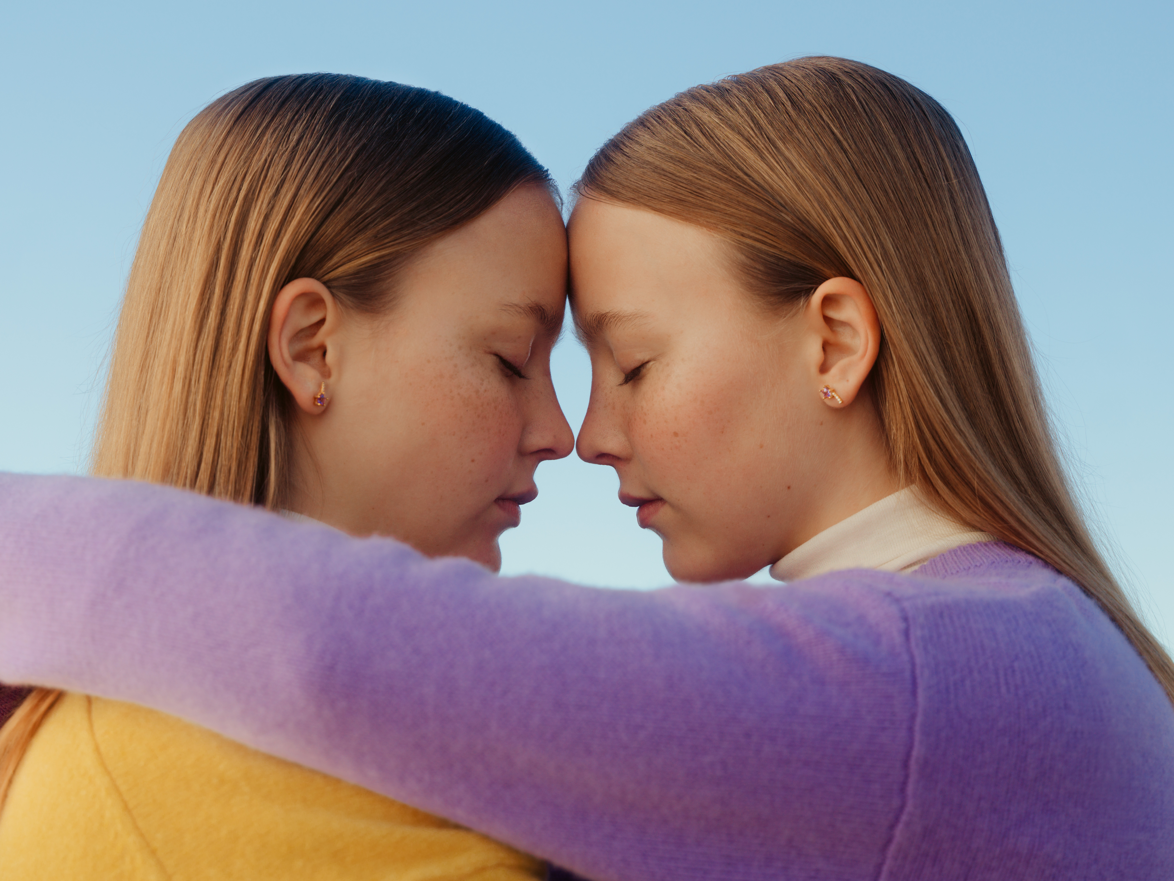 Image of two young women in orange and purple sweaters from the side. They hold their heads together and their foreheads are touching.