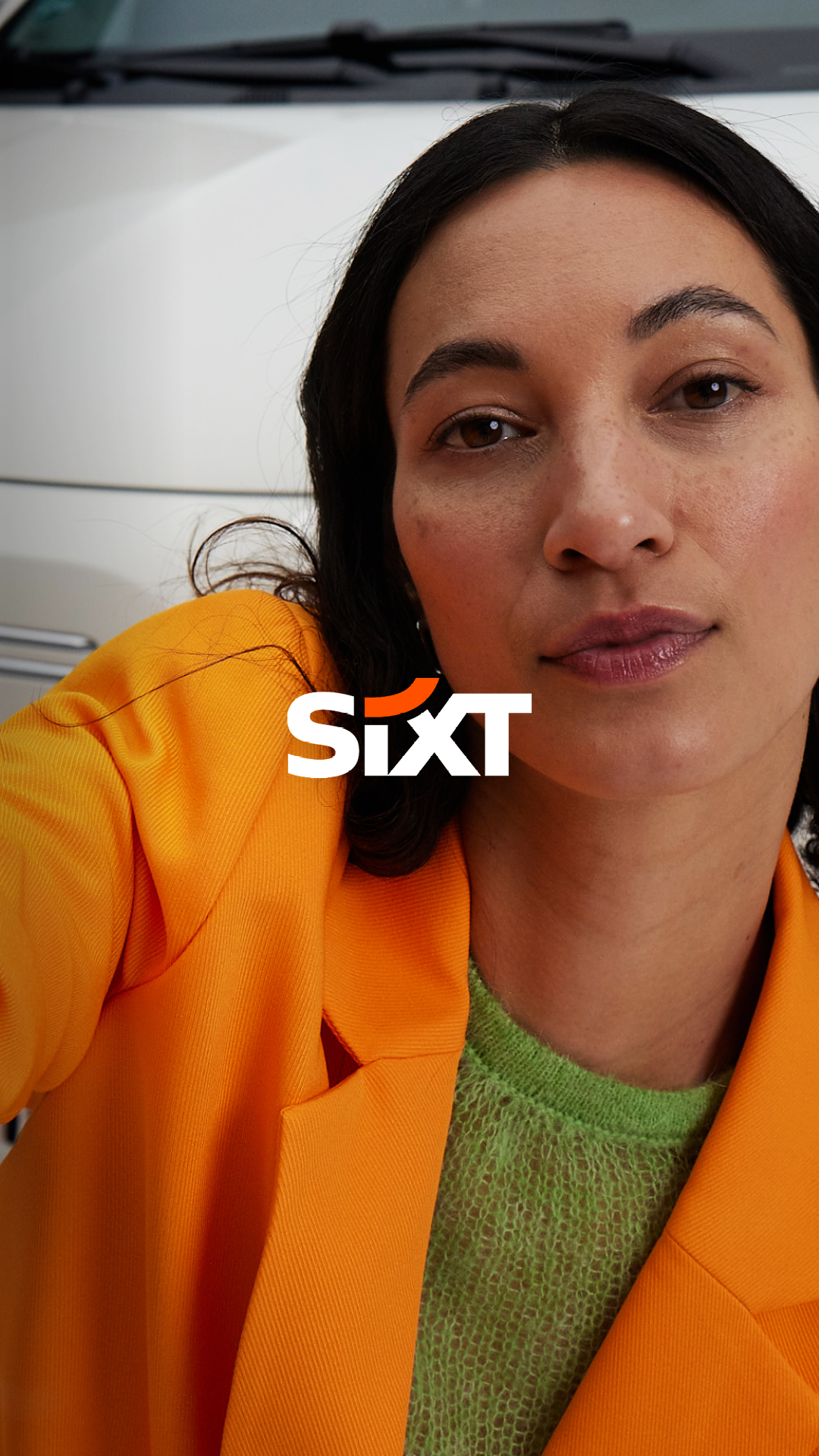 SIXT ad in new brand design by Jung von Matt BRAND IDENTITY: a young women with a blazer jacket in SIXT Orange standing in front of a white FIAT 500 making a selfie with her smartphone, the hand a bit in front of the camera