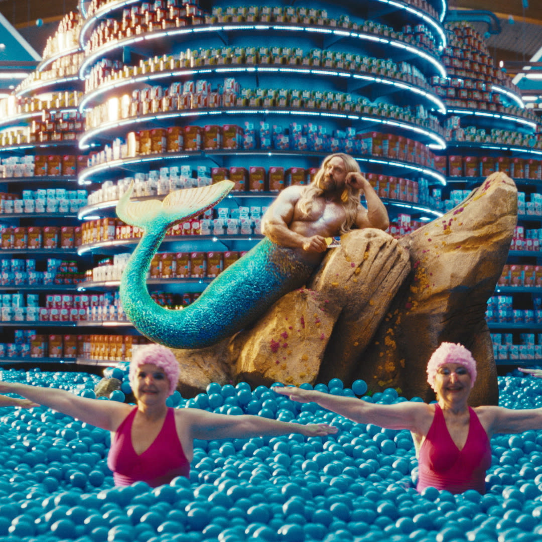 A whimsical scene in a supermarket featuring multiple women in pink swimsuits and swim caps standing in blue plastic balls. In the background, a large sculpture of a mermaid and a man lounges on a rock, surrounded by shelves filled with colorful products.