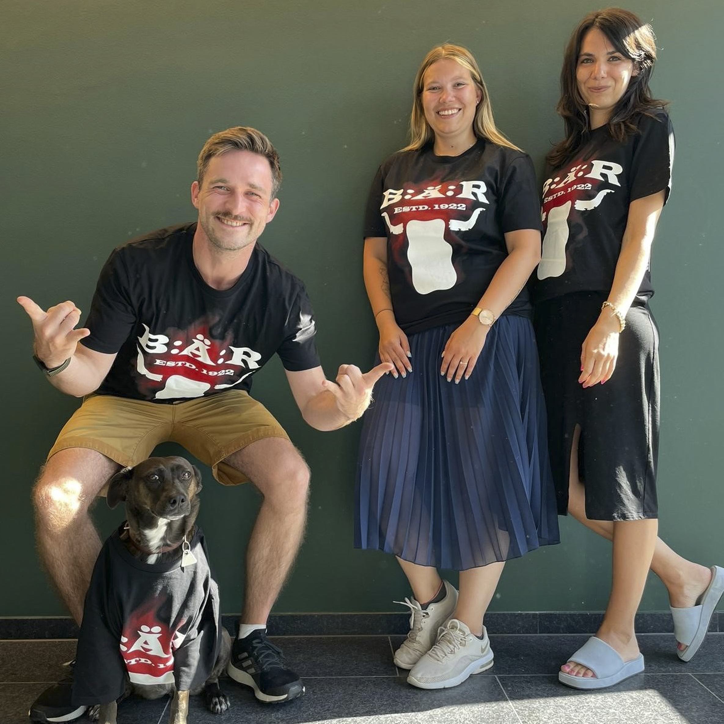 Picture of three employees of the Jung von Matt Netzeffekt office in Munich. All are smiling and wearing shirts from the HARIBO and Wacken collaboration campaign. A dog wearing the same shirt is sitting at the bottom left of the picture.