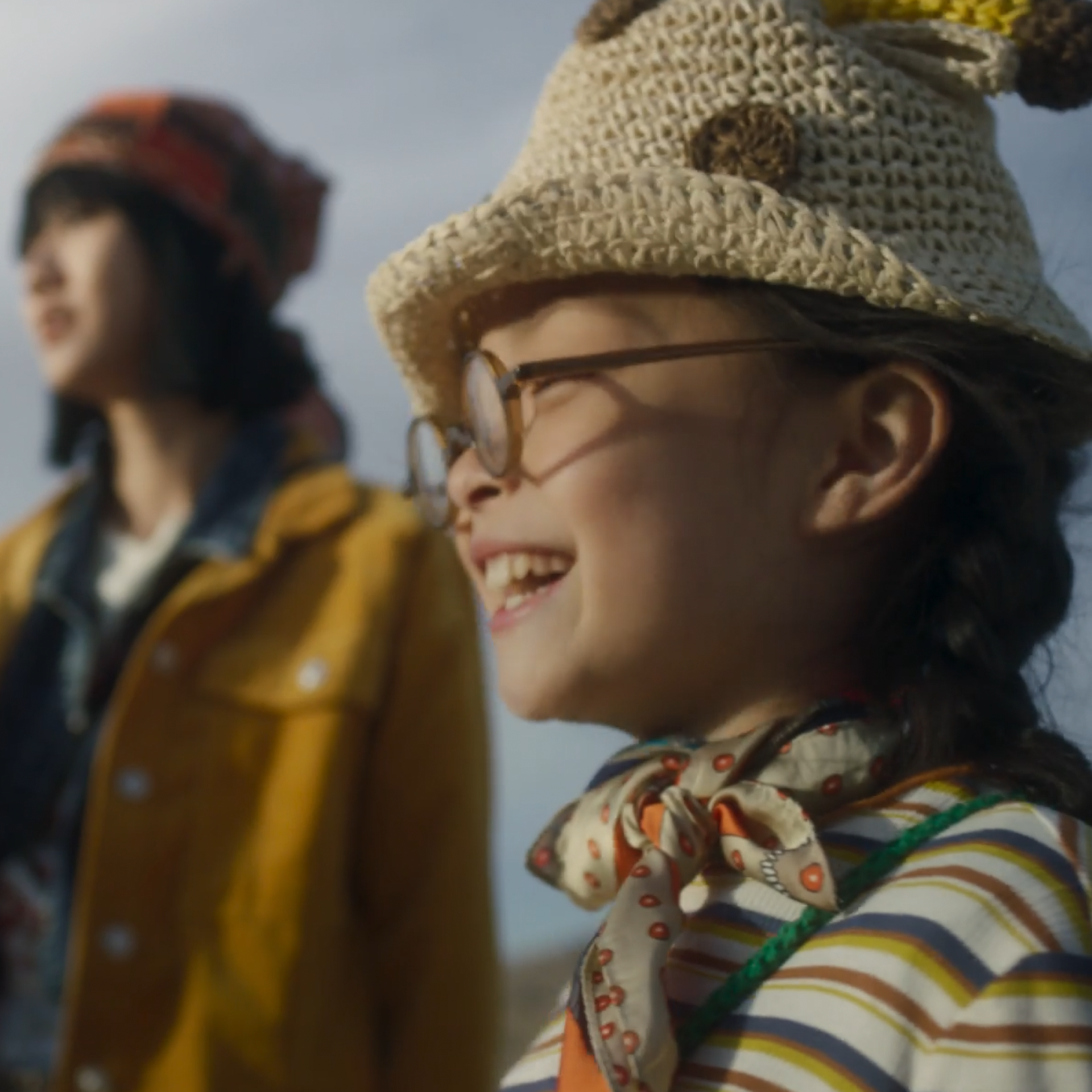 Image of a little smiling girl with glasses wearing a cap. You can see other people in the background.
