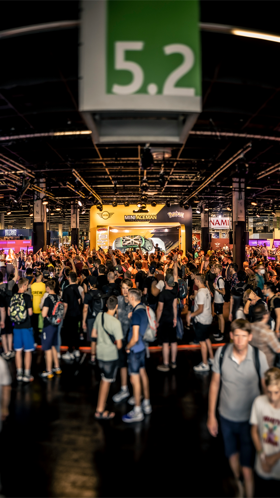 A crowd of people walking through an exhibition hall at gamescom 2022. In the background, the concept car Aceman in life-size toy packaging can be seen.