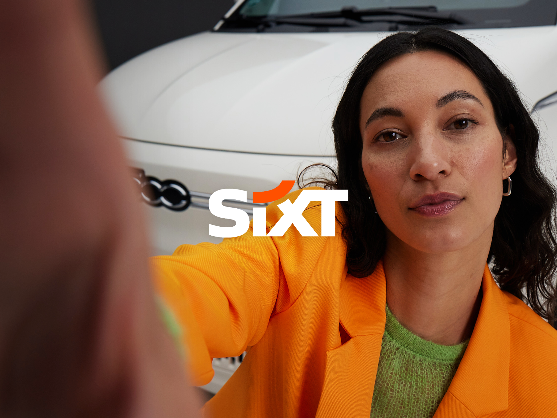 Image of a woman in orange blazer taking a selfie of herself. In the background you can see a white Fiat 500. In the middle as an overlay the slogan "Sixt".