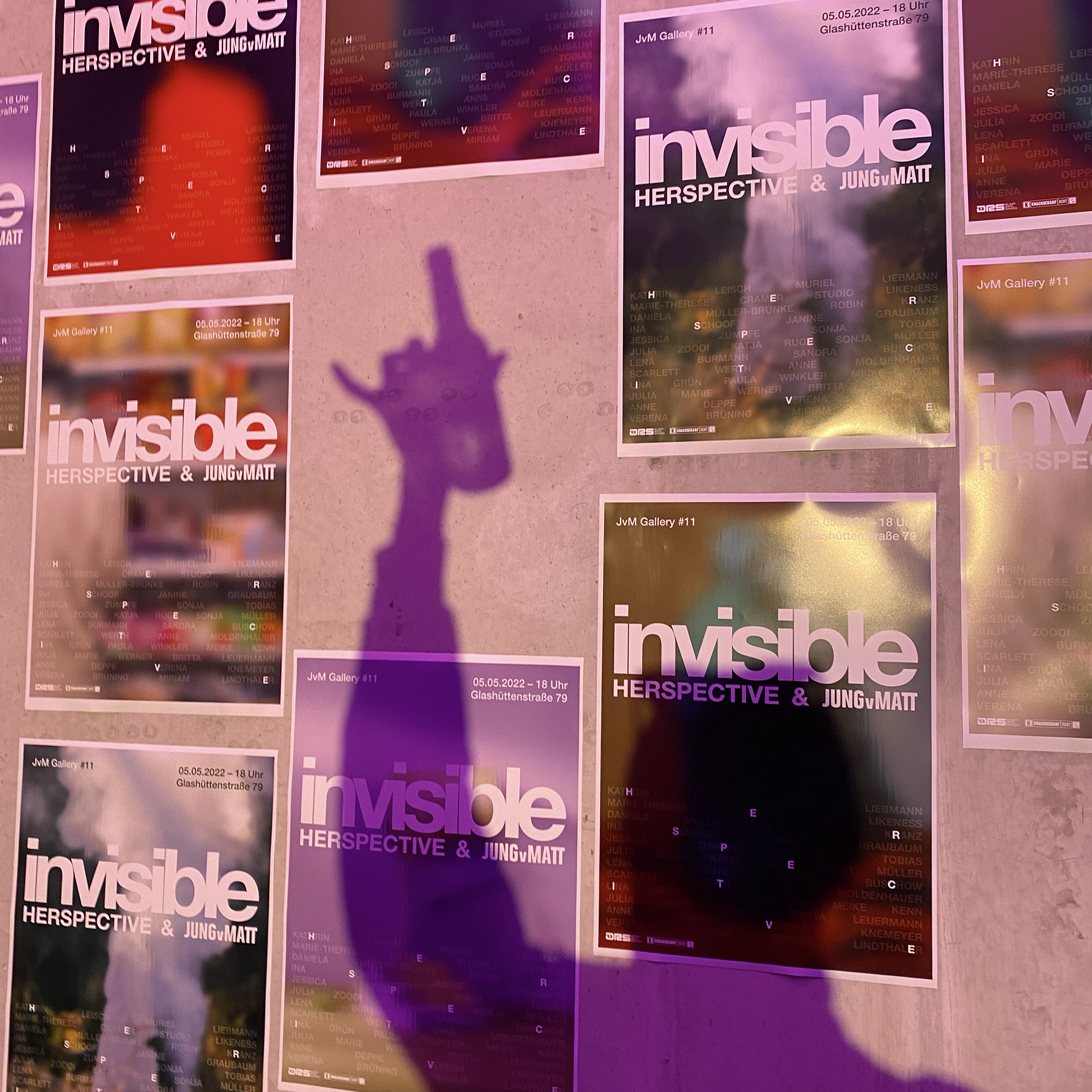 Image of a wall on which posters of the of "Invisible. Herspective & Jung von Matt" can be seen. A held up arm with a drink in the hand casts a shadow.