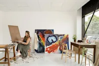 An atelier with a desk, finished paintings and a woman painting next to an easel and table