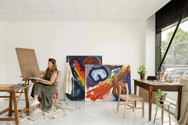 An atelier with a desk, finished paintings and a woman painting next to an easel and table
