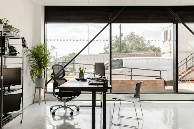 An office with a desk, a shelf and a big plant with three large windows overlooking a street with a van
