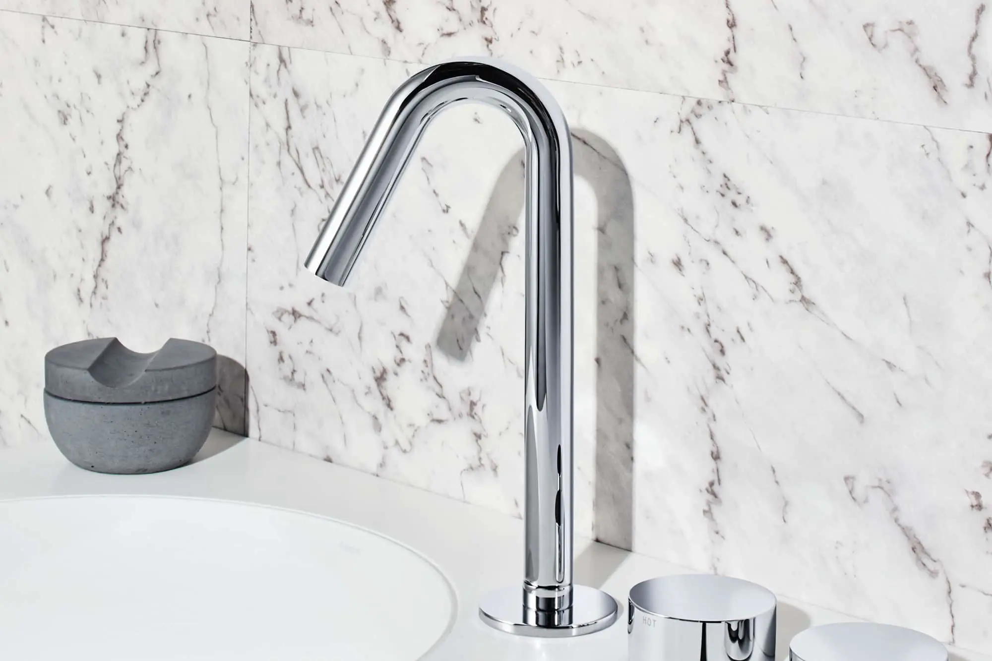 Chrome bathroom faucet with curved spout and separate handles on white marble countertop with gray soap dish