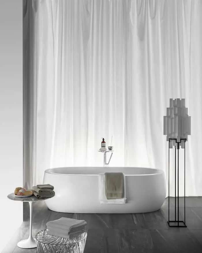 Modern white freestanding bathtub with towels, side table, and geometric floor lamp against white curtains.