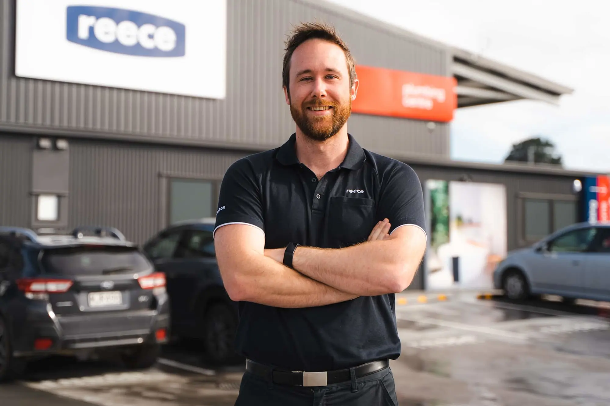 Man in black polo shirt with arms crossed standing in front of a building with Reece signage and parked cars.