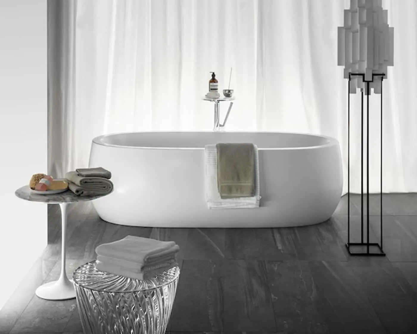 White freestanding bathtub with towels and bath accessories in a minimalist bathroom with white curtains and gray floor.