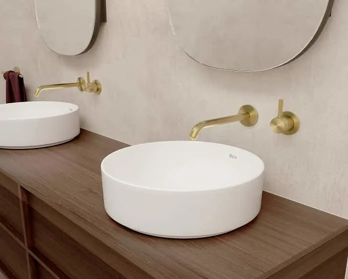 Modern bathroom with two white round vessel sinks on wood counter, brass wall-mounted faucets, and round mirrors.