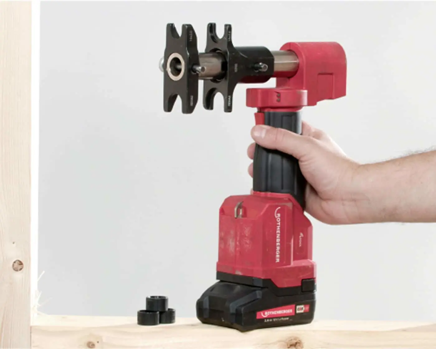 Hand holding a red cordless pressing tool with black attachment against a white wall near wooden surface.