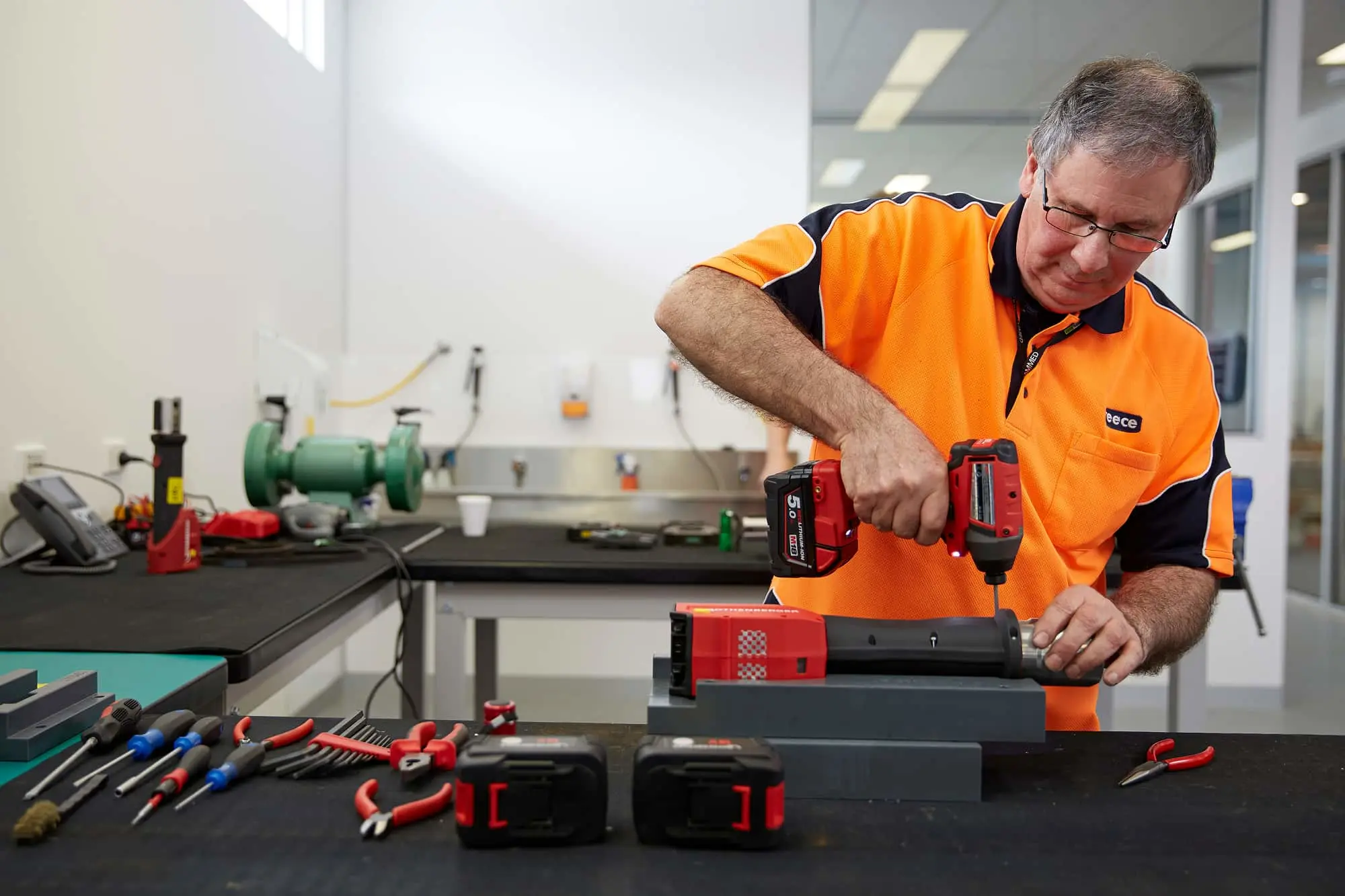 Man in orange safety vest using power drill on equipment at workbench with tools in workshop.