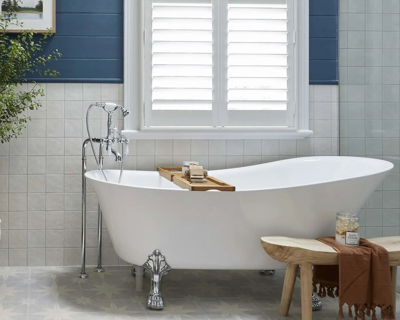 White freestanding bathtub with chrome faucet in tiled bathroom with white shuttered window and wooden bath caddy.