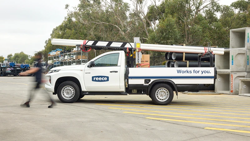 White Reeco utility truck with roof rack and pipes parked in outdoor lot with person walking past and trees behind.