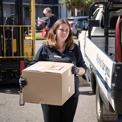 Woman in dark uniform carrying cardboard box near delivery truck outdoors on sunny day.