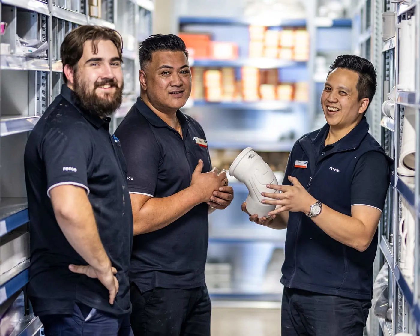 Three workers in navy uniforms standing in a warehouse aisle with shelving, two holding a white container.