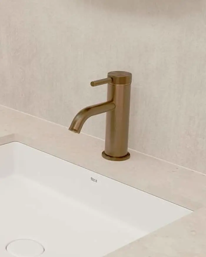 Brushed gold bathroom faucet on white countertop with rectangular sink against beige tile wall