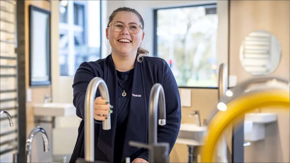 Woman wearing glasses and black uniform standing next to chrome faucet displays in a showroom with windows in background.