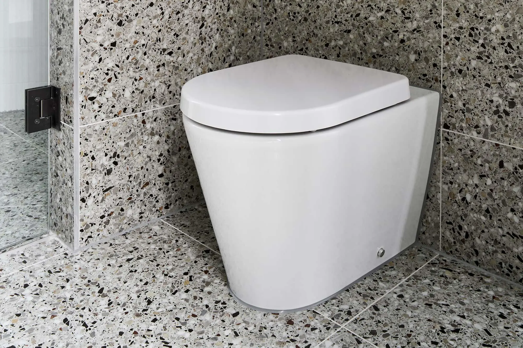 White modern toilet in corner of bathroom with terrazzo tile walls and floor featuring black and beige speckles.