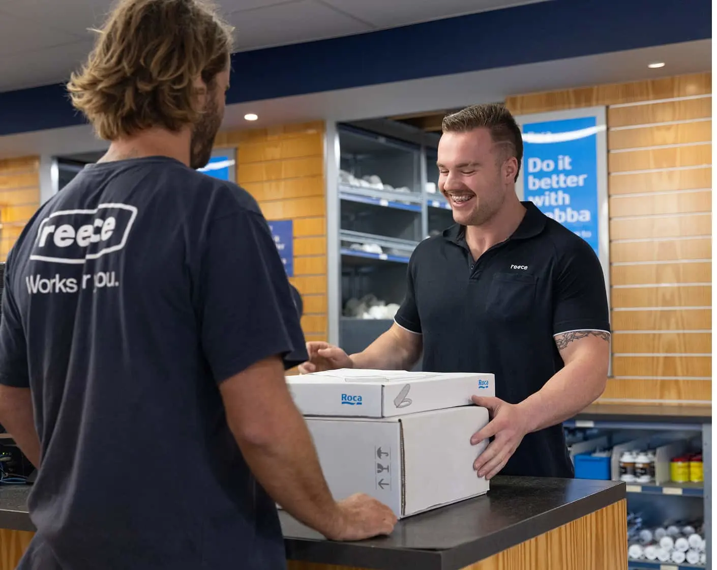 Two men in black polo shirts at a counter, one handing a white Roca product box to the other in a retail setting.