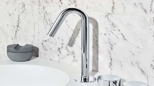 Chrome bathroom faucet with curved spout and separate handles on white marble countertop with sink and gray soap dish