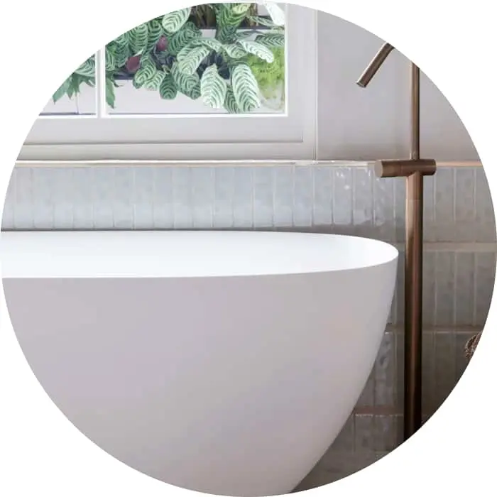 White freestanding bathtub with bronze floor-mounted faucet against white tiled wall beneath window with plant