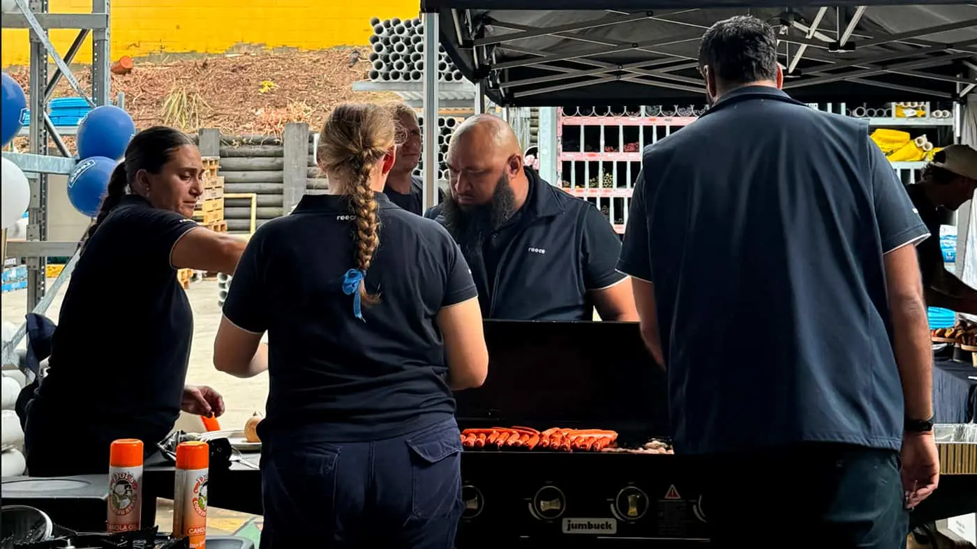 Four people in dark polo shirts grilling sausages outdoors under a black canopy with blue balloons visible.